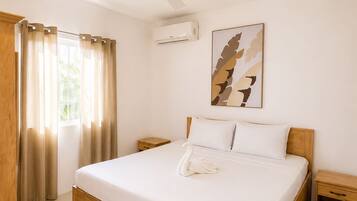 Habitación de hotel con cama de madera, ventilador de techo, unidad de aire acondicionado, ventana con cortinas y una obra de arte colgada en la pared.