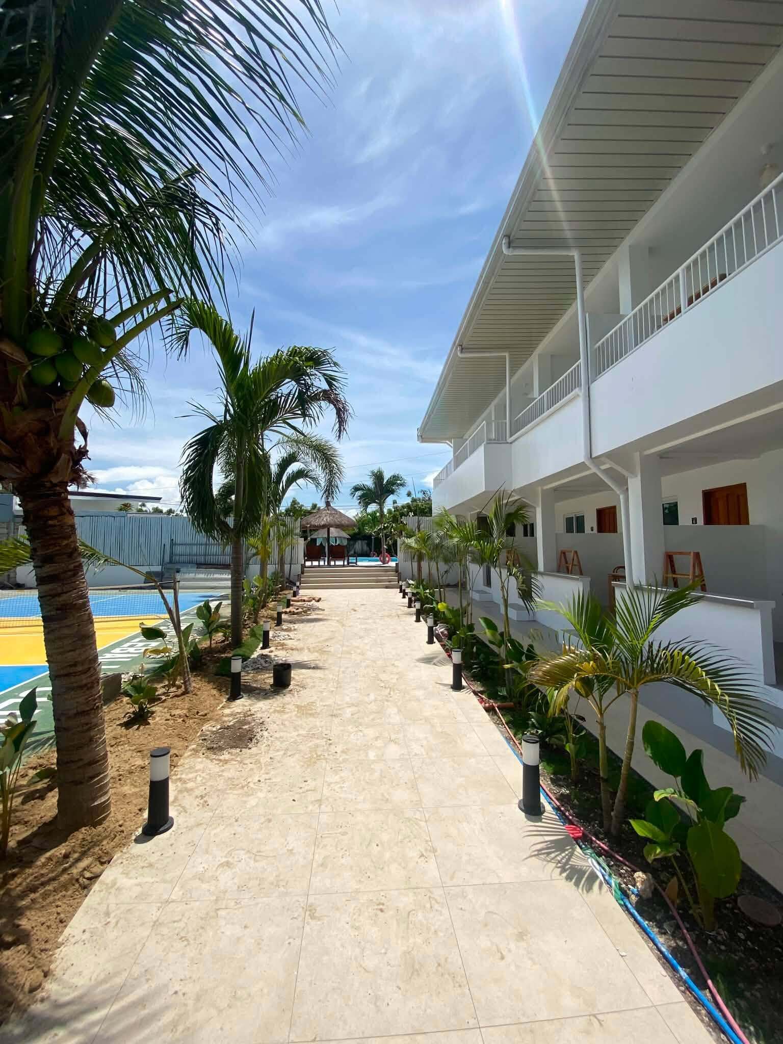 A pathway lined with palm trees leading to a pool area, with a white building featuring balconies and a covered seating area in the background.