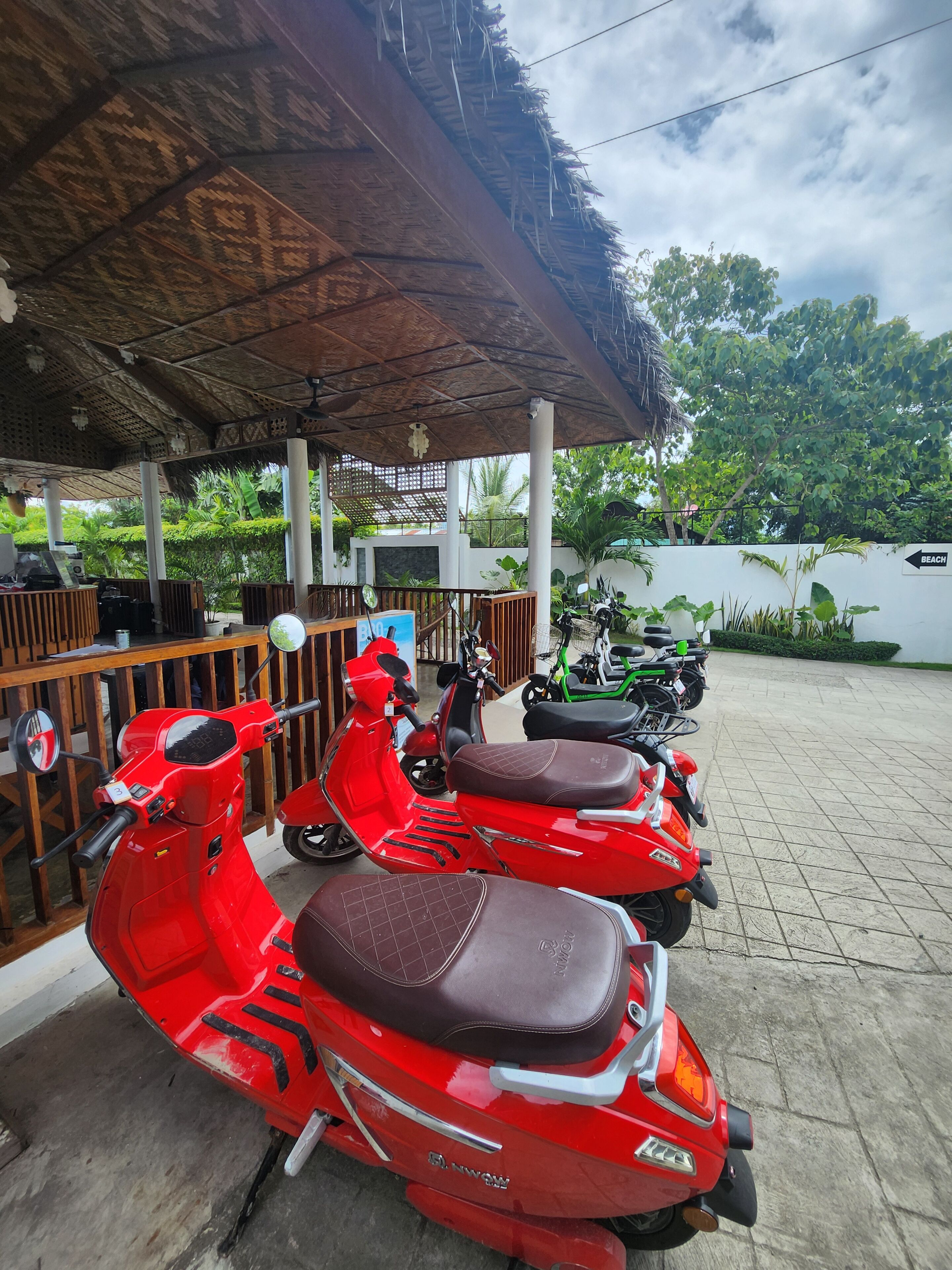 Un espacio de estar al aire libre techado con techo de paja, una fila de scooters rojos y un cartel que indica la dirección hacia la playa.