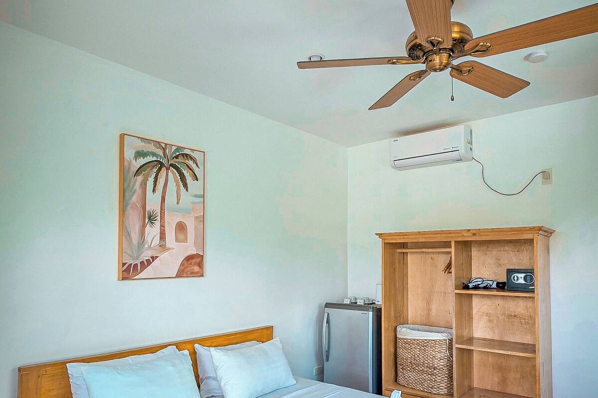 A bedroom with a bed, a ceiling fan, a wall-mounted air conditioner, a television, and a wooden cabinet.