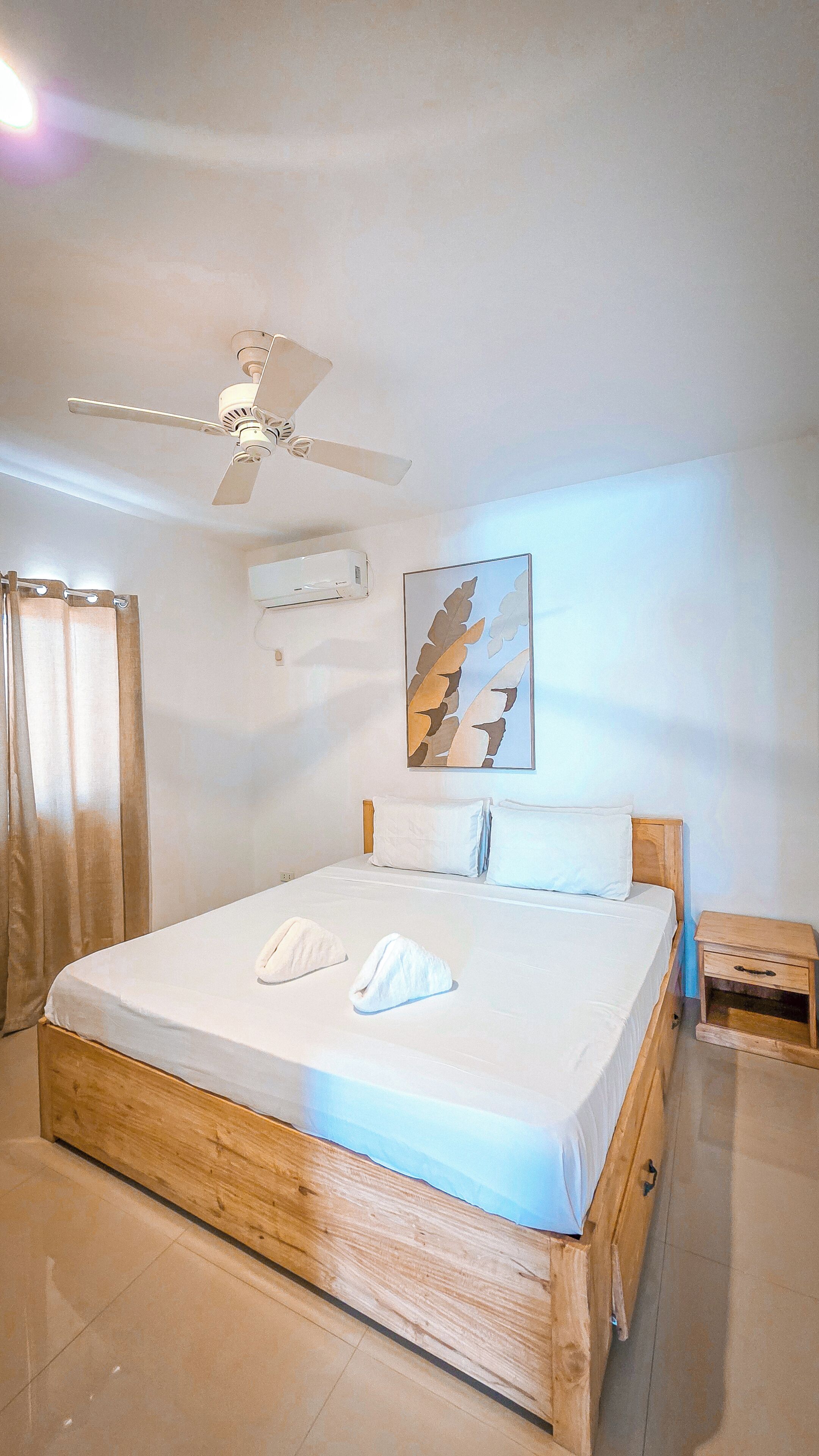 A hotel room with a bed, a ceiling fan, a painting, and a wooden nightstand.