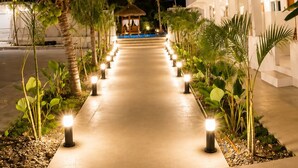 A pathway lined with palm trees and illuminated bollards leading to a building with balconies.