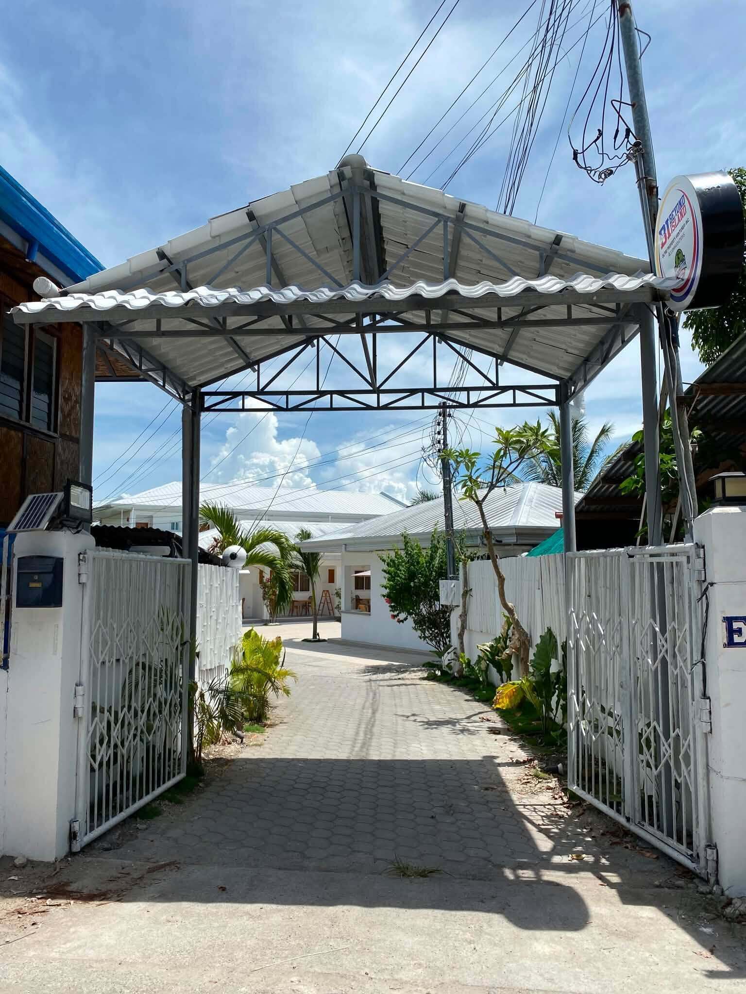 A gated entrance with a metal roof, a paved pathway, and a building with a white exterior and a blue roof in the background.