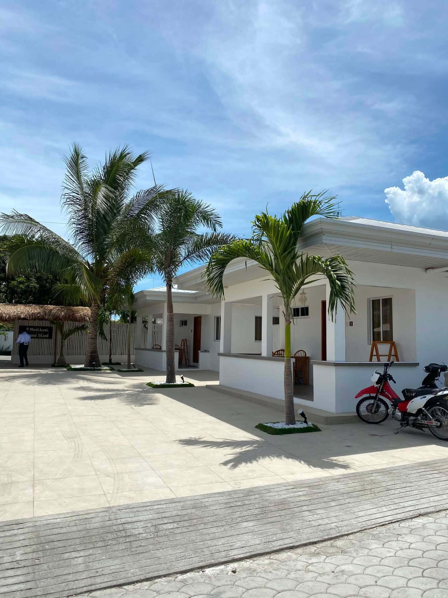 A hotel courtyard with palm trees, a red motorcycle, and a thatched-roof structure.