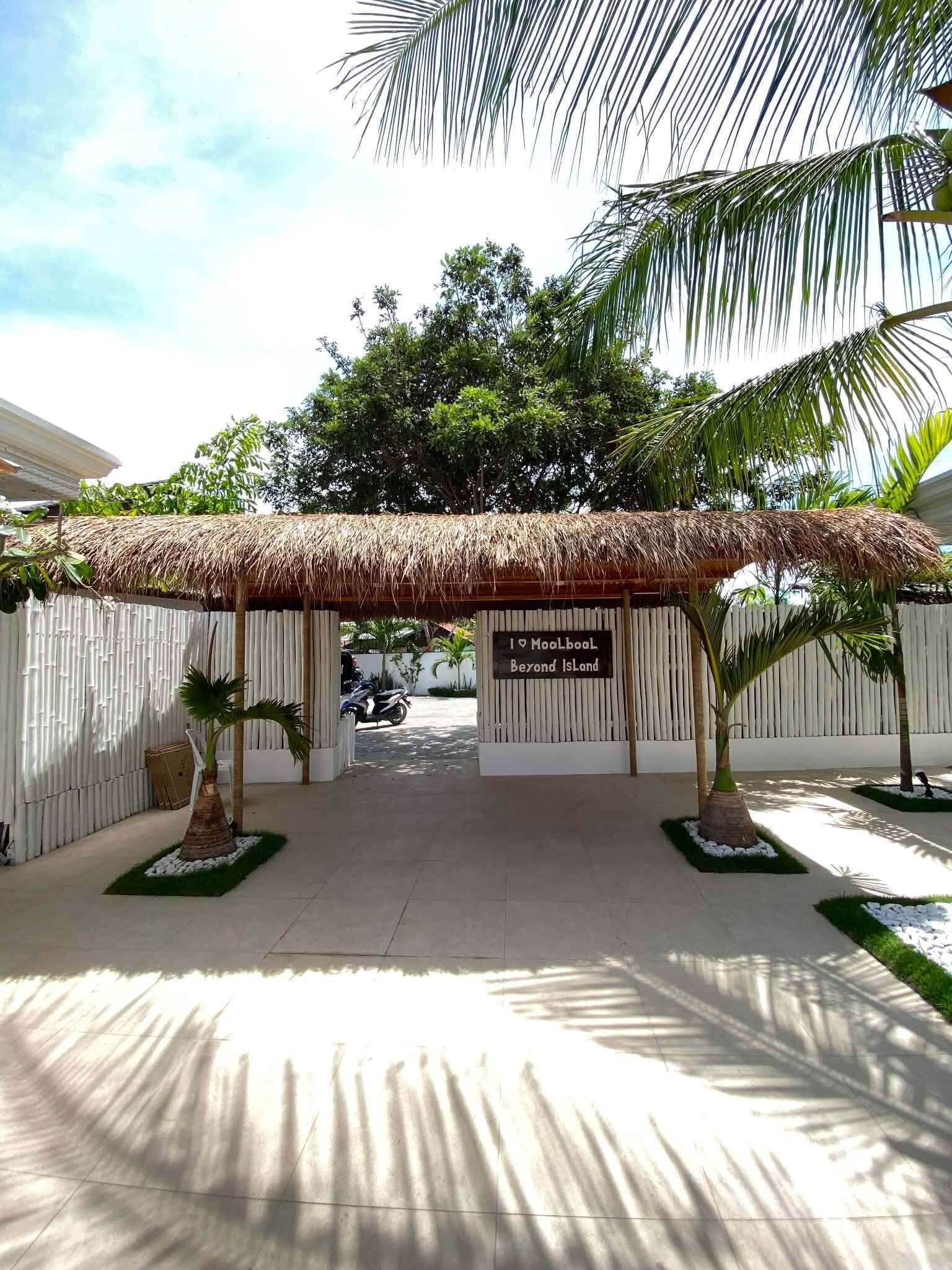 A covered entrance with a sign, palm trees, and a white fence.