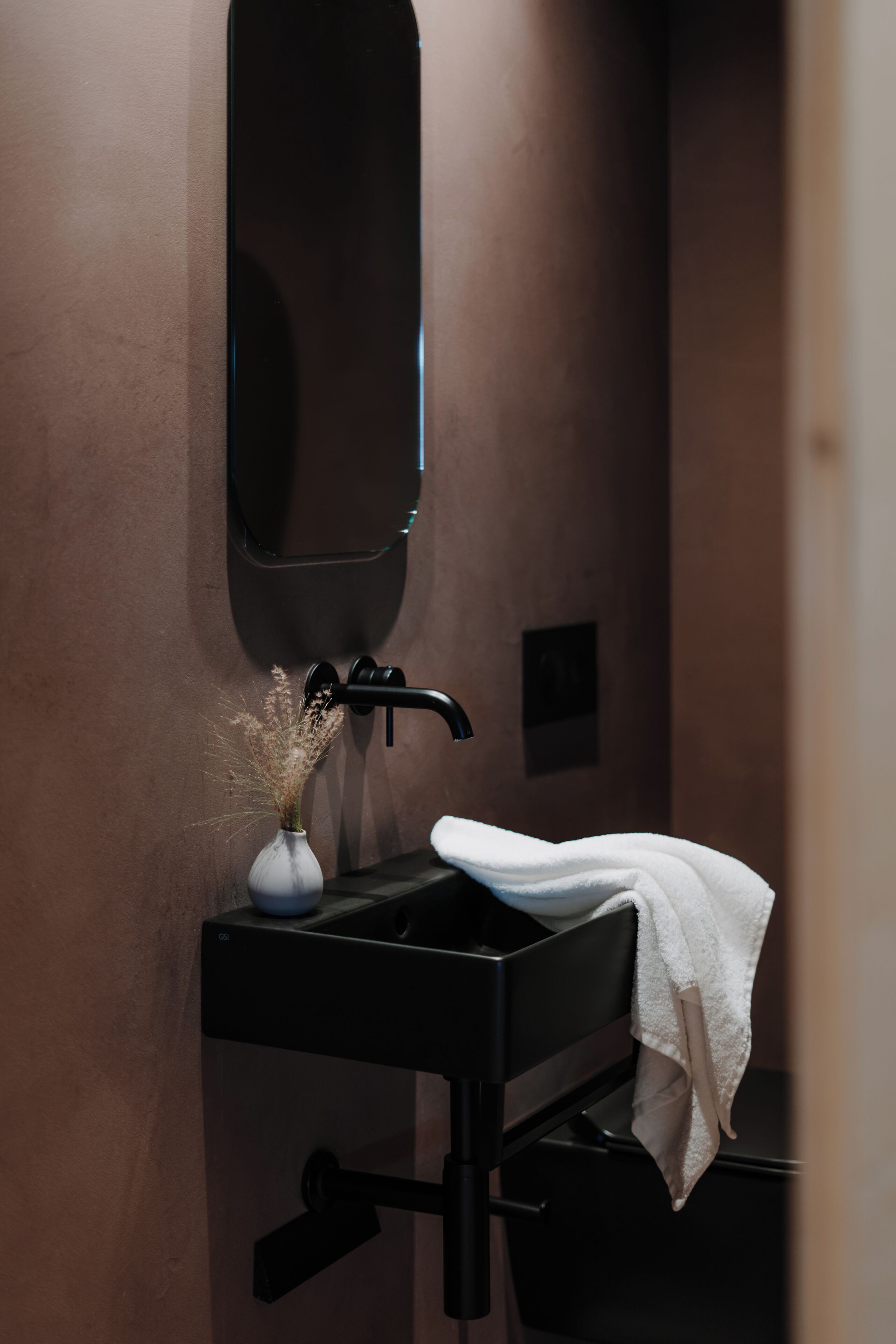 Un baño con un lavamanos negro, un espejo, un florero con flores secas y una toalla.