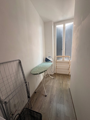 Una habitación pequeña con suelo de madera, un estante de metal, un tendedero de madera y una ventana con persianas.