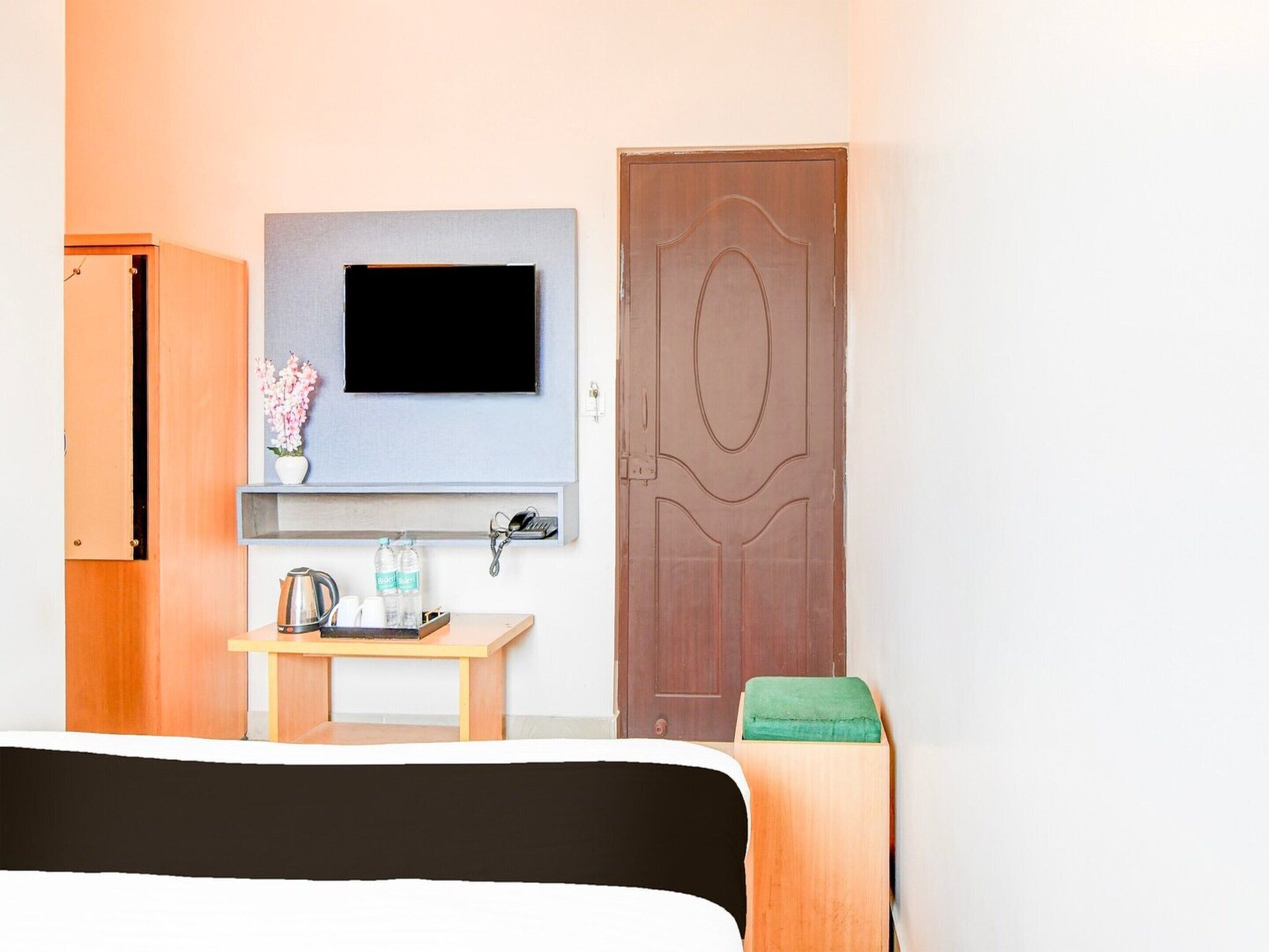 A hotel room with a bed, a TV mounted on the wall, a small table with a kettle and bottles, a wooden door, and a green cushion.