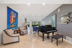 A spacious living room with a grand piano, comfortable seating, a large poster, and a collection of books on the wall. - Courtside Escape - Country Club Living (Rancho Mirage)