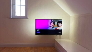 Une pièce avec un téléviseur fixé au mur, une fenêtre, un tapis à motifs et un plancher en bois.