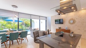 Eine moderne Küche mit einer großen Kochinsel, einem Essbereich mit Stühlen, einem an der Wand befestigten Fernseher und Blick ins Grüne.