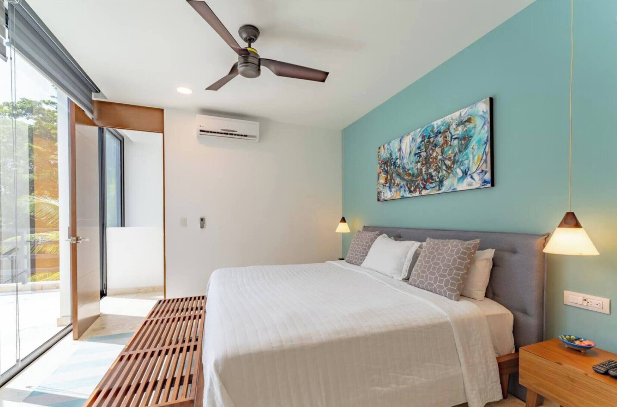 Un dormitorio moderno con cama, ventilador de techo, obras de arte y vistas a través de un ventanal.