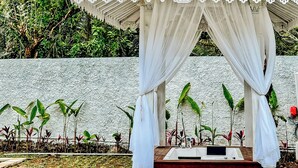 A covered outdoor area with a wooden platform, white drapes, and potted plants against a textured wall. - Hafrana Villa by AGATA - 700sqm 2BR Villa with Private Pool & Jacuzzi (Selat)