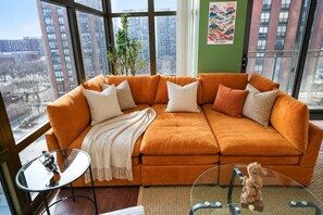A modern living room with a large orange sectional sofa, a glass coffee table, and a view of a cityscape through large windows.