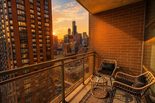 A balcony with wicker chairs and a view of a cityscape at sunset.