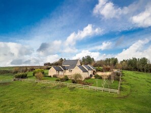 A stone farmhouse with multiple attached buildings, surrounded by green fields and a wooden fence.