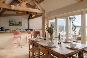 A dining area with a wooden table set for four, a kitchen in the background, and a view of the outside.
