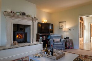 A living room with a fireplace, a television, a blue armchair, and a table with a tray of wine glasses and cookies.