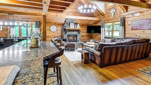 A log cabin interior with a stone fireplace, wooden ceiling beams, a bar area, and a leather sofa. - Hot Tub & Patio: Bedford Log Cabin on 10 Acres! (Bedford)