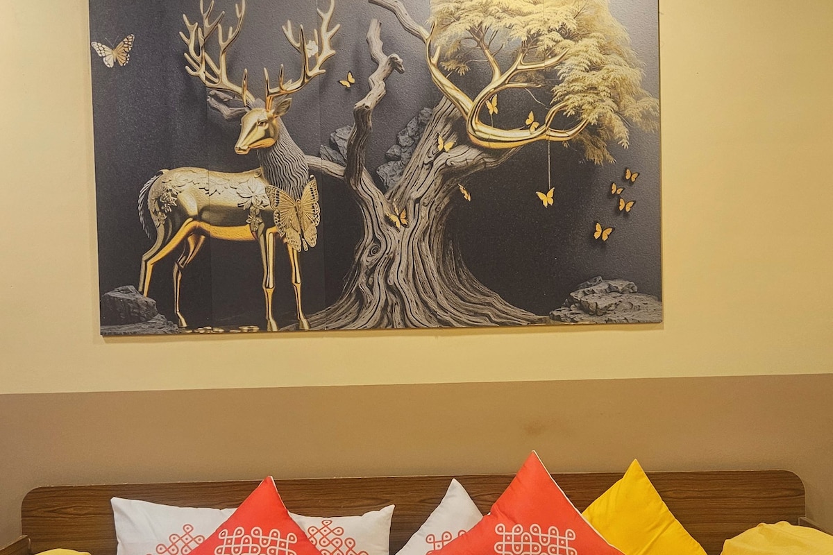 A bed with a yellow bedspread, a wall-mounted painting of a deer and tree, and patterned pillows.