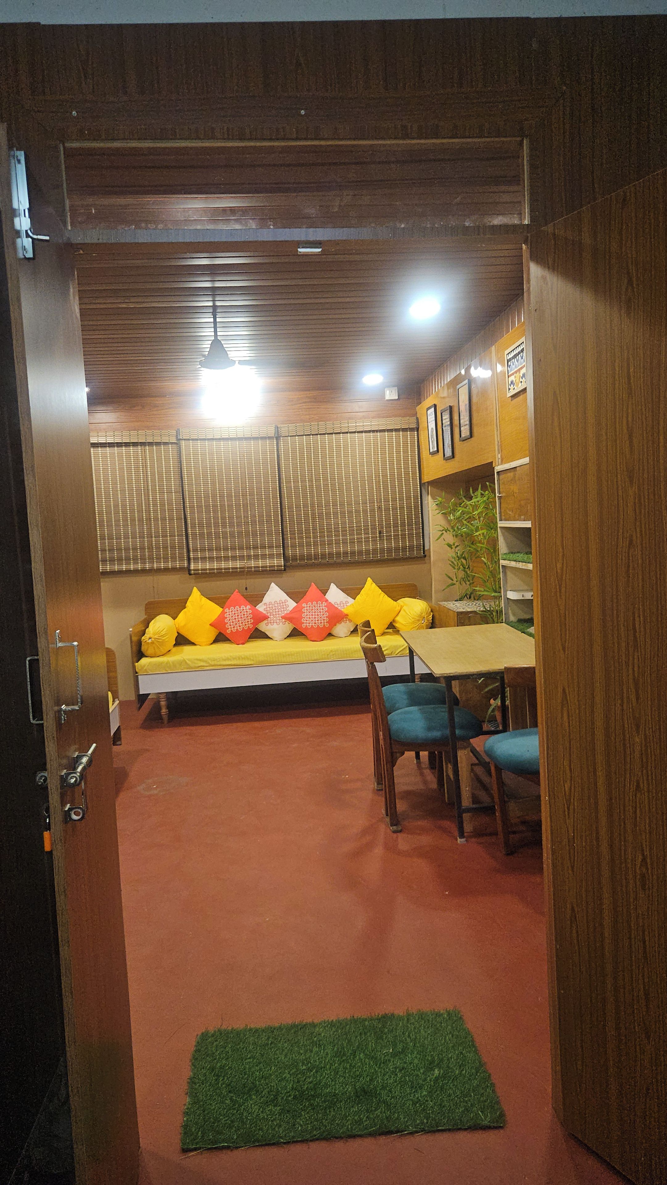 A room with a wooden ceiling, a sofa with cushions, a table, chairs, and a green rug.