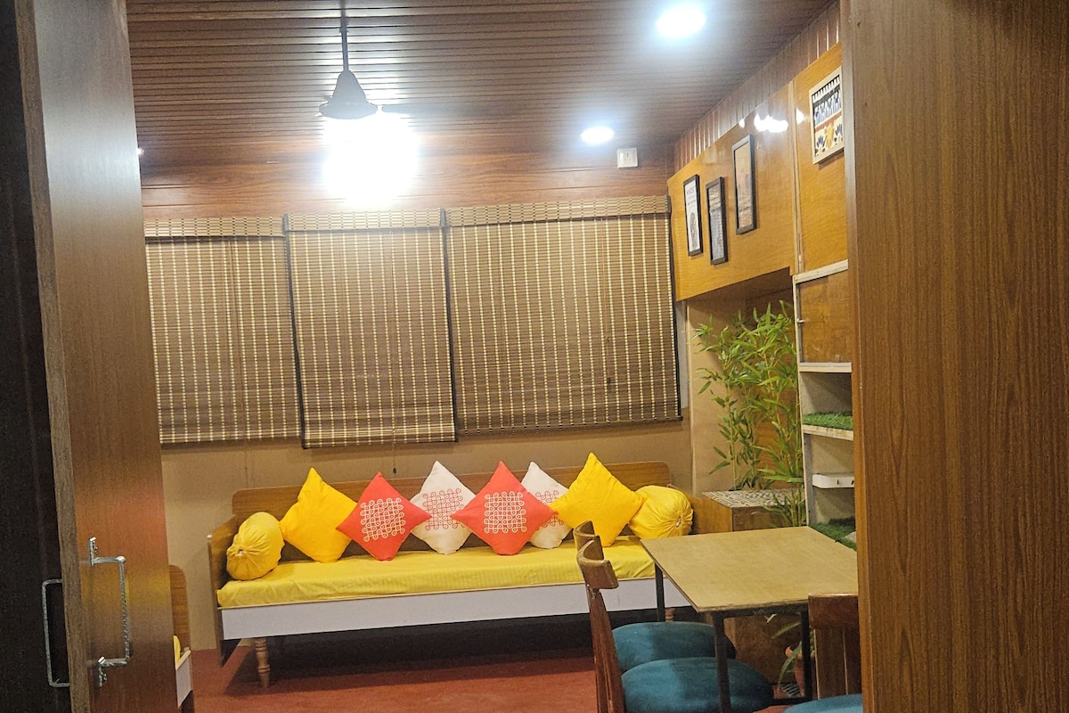 A room with a wooden ceiling, a sofa with cushions, a table, chairs, and a green rug.
