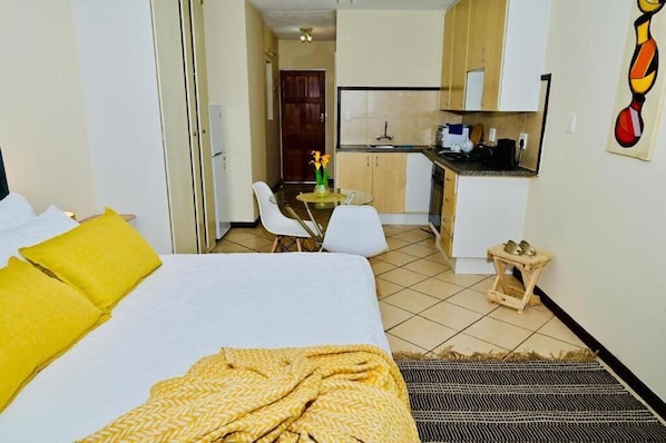 Una habitación de hotel con una cama, una pequeña mesa con sillas, una kitchenette y una puerta que da a otra habitación.