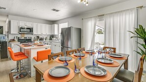 A modern kitchen with white cabinets, a stainless steel refrigerator, and a dining area with orange chairs and a wooden table set for six. - Outdoor Oasis & All-Ages Fun! Gulf Coast Getaway (Hudson)