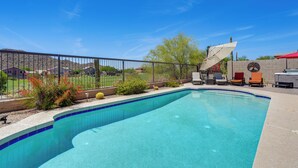 Ein Pool mit geschwungener Kante, umgeben von einem Zaun, und eine Terrasse mit Stühlen und einem Grill.