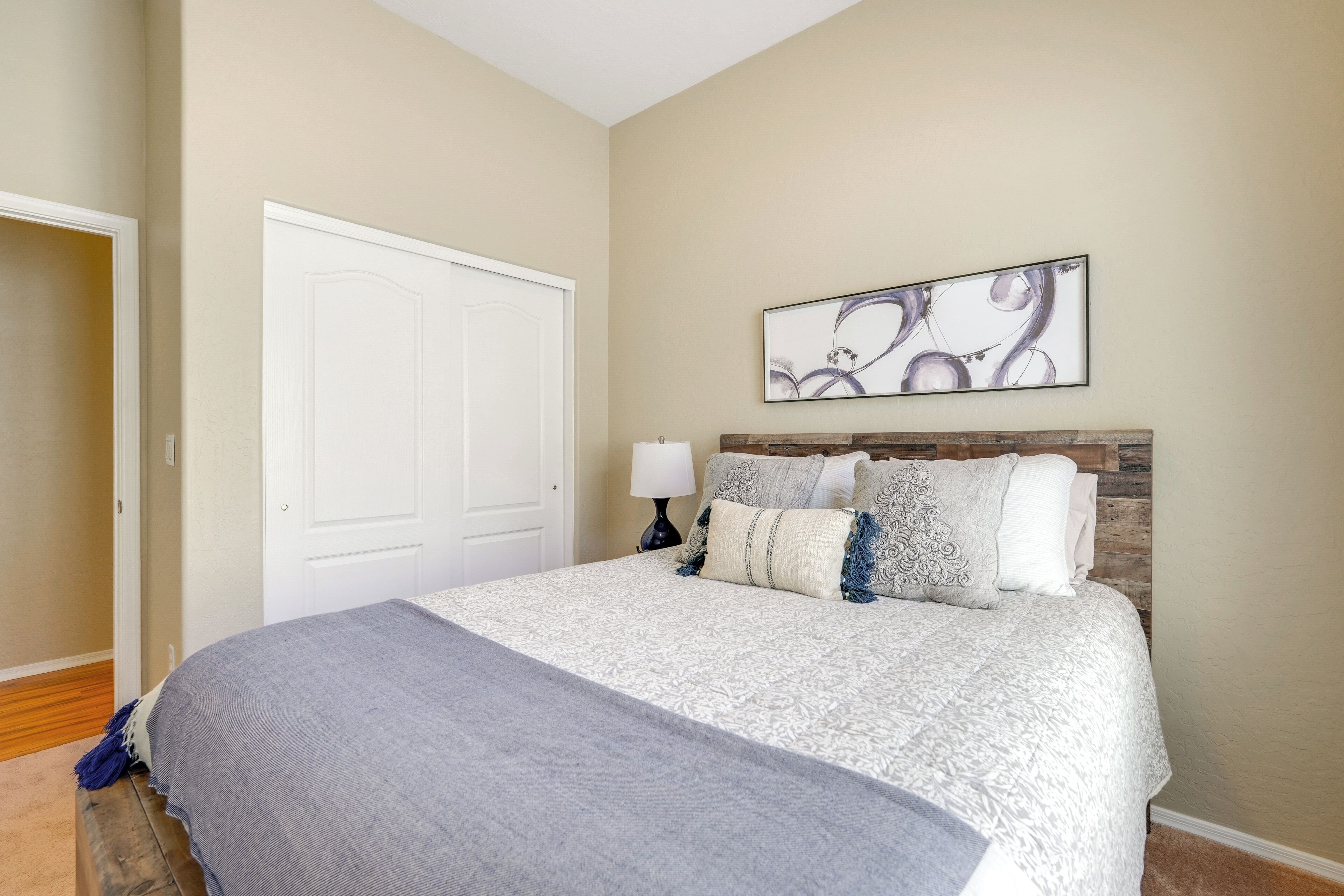 Un dormitorio con cama, puerta, una obra de arte en la pared y una mesita de noche con lámpara.