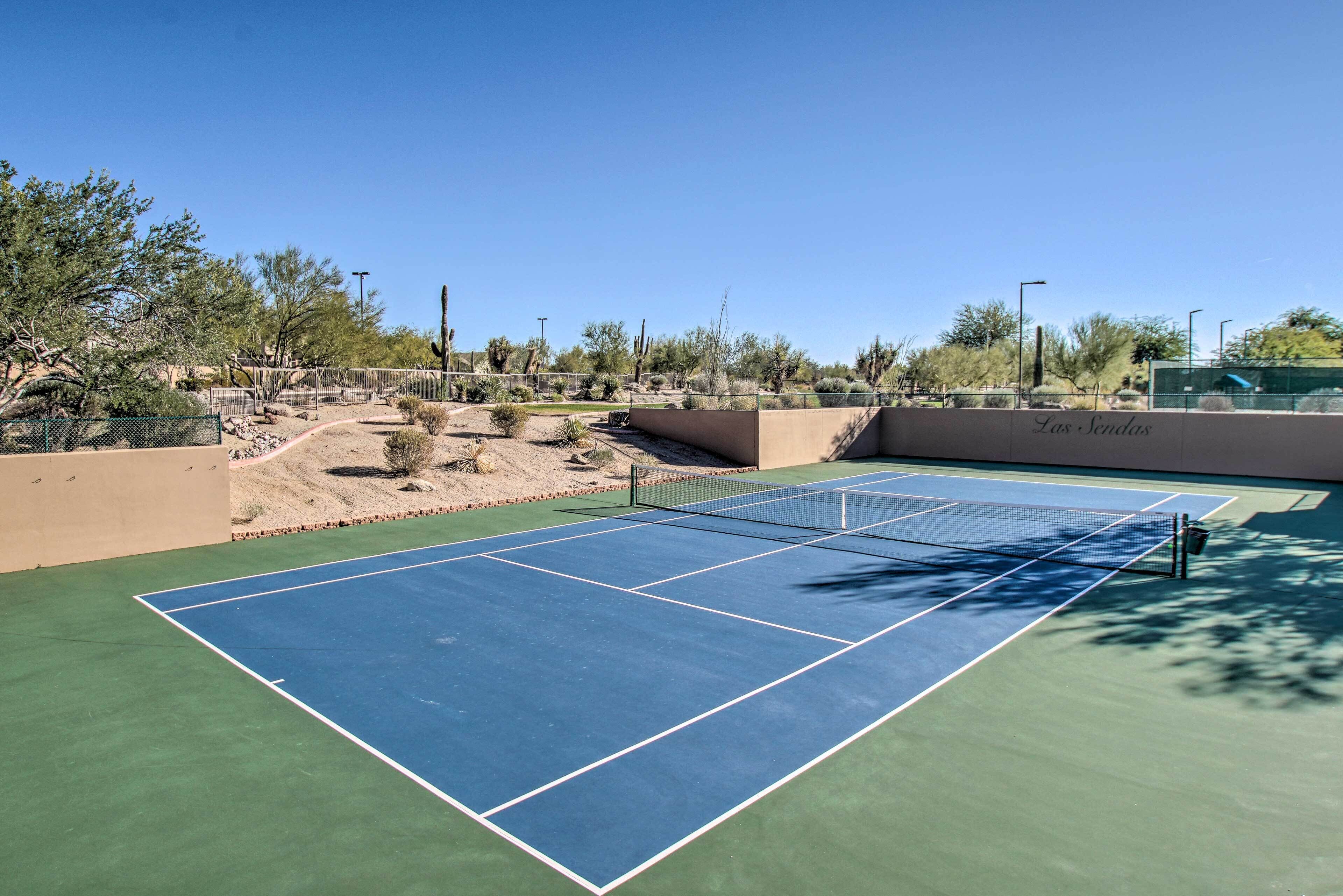 Una cancha de tenis al aire libre con cielo despejado, rodeada de vegetación desértica y una valla.