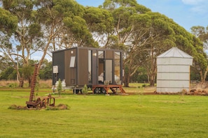 Une petite cabane dotĂ©e dâune porte coulissante, un abri sĂ©parĂ© servant de remise, et une structure mĂ©tallique rouillĂ©e, le tout situĂ© dans un champ herbeux avec des arbres en arriĂšre-plan.