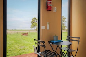 A room with a view of a grassy field, a fire extinguisher, a thermostat, and a table with chairs. - Lara Grove Cottage 1 by Tiny Away (Purdeet)