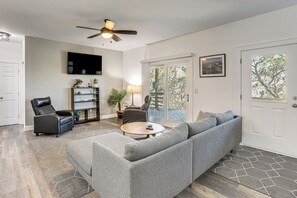 A living room with a gray sectional sofa, a round coffee table, a leather chair, a TV mounted on the wall, and a ceiling fan.