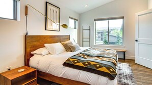 Een slaapkamer met een houten hoofdbord, een bed met een gebedrukte deken, een nachtkastje en een raam met uitzicht op een gebouw en bomen.