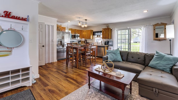 A spacious living room with a sectional sofa, a dining area with wooden chairs, a kitchen with wooden cabinets, and a large window. - Dog-Friendly Waneta Lake Home w/ Lake Access! (Wayne)