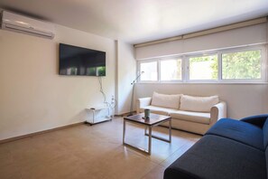 A living room with a flat-screen TV, a white sofa, a blue sofa, a glass-top coffee table, and a window with blinds. - Elegant Urban Retreat in Psychiko 2 (Psichiko)