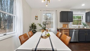 A dining area with a table set for two, a kitchen with dark cabinets, and a view of trees through the windows. - Catskill Chalet by Avantstay Sleeps 22 Hot Tub + Firepit + Playroom Mins to Hunter Mtn (Tannersville)