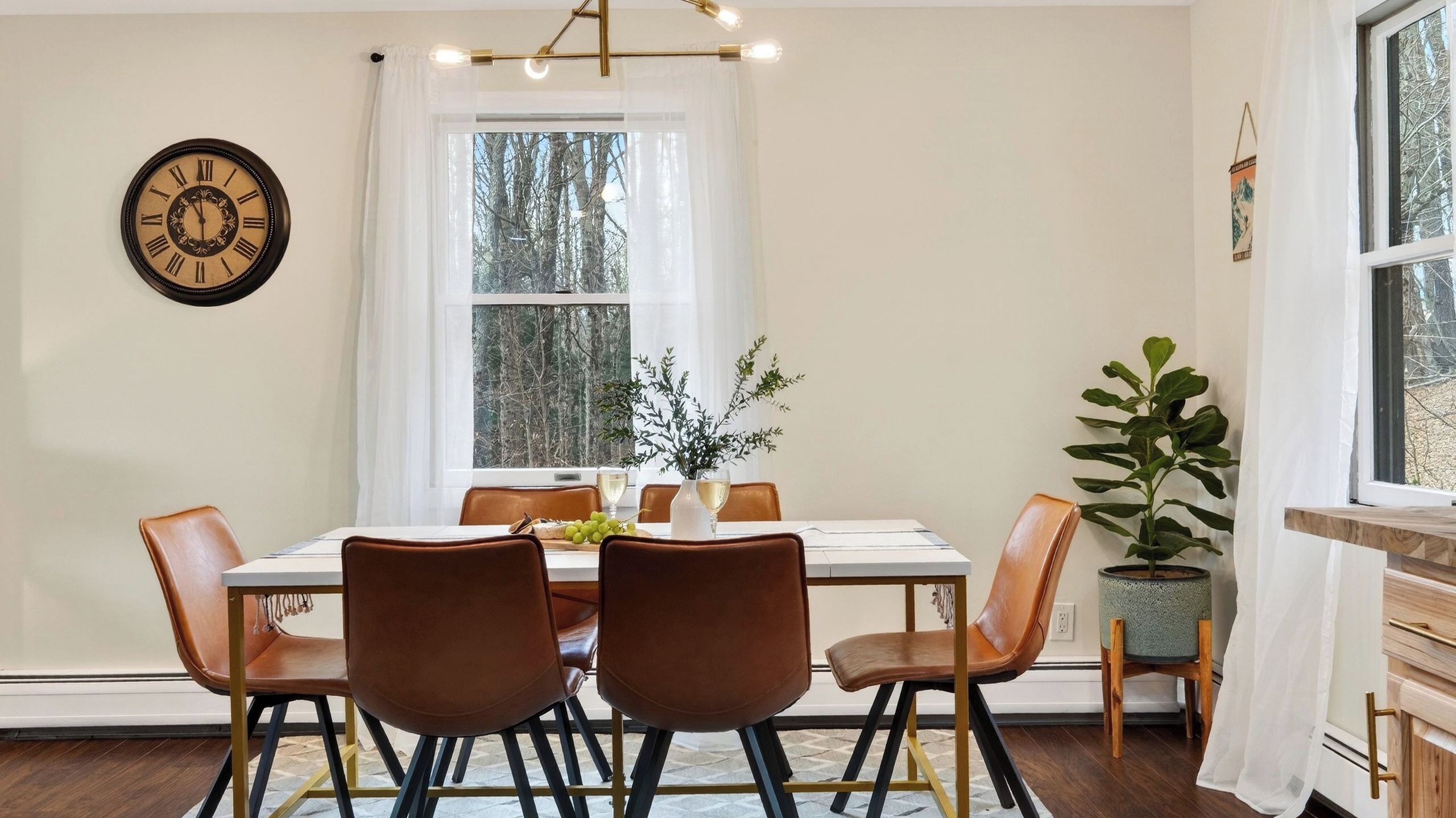 Un comedor con una mesa, sillas, un reloj y una ventana con vistas a árboles.