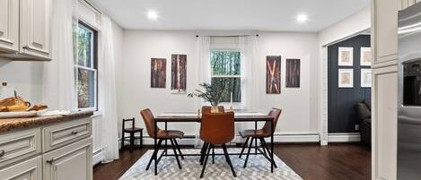 A kitchen with a dining area, white cabinetry, a refrigerator, and a window with a view of trees.