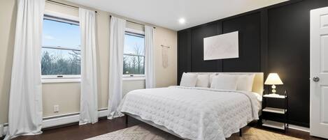 A bedroom with a bed, a nightstand, a lamp, a wall-mounted artwork, and a window with curtains.