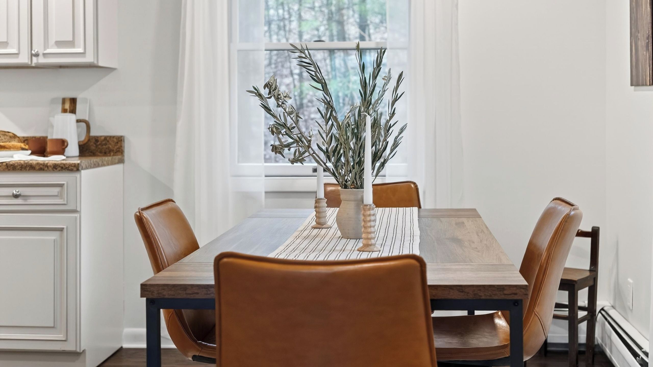 Une table à manger avec des chaises, un vase avec des branches et une fenêtre avec des rideaux.