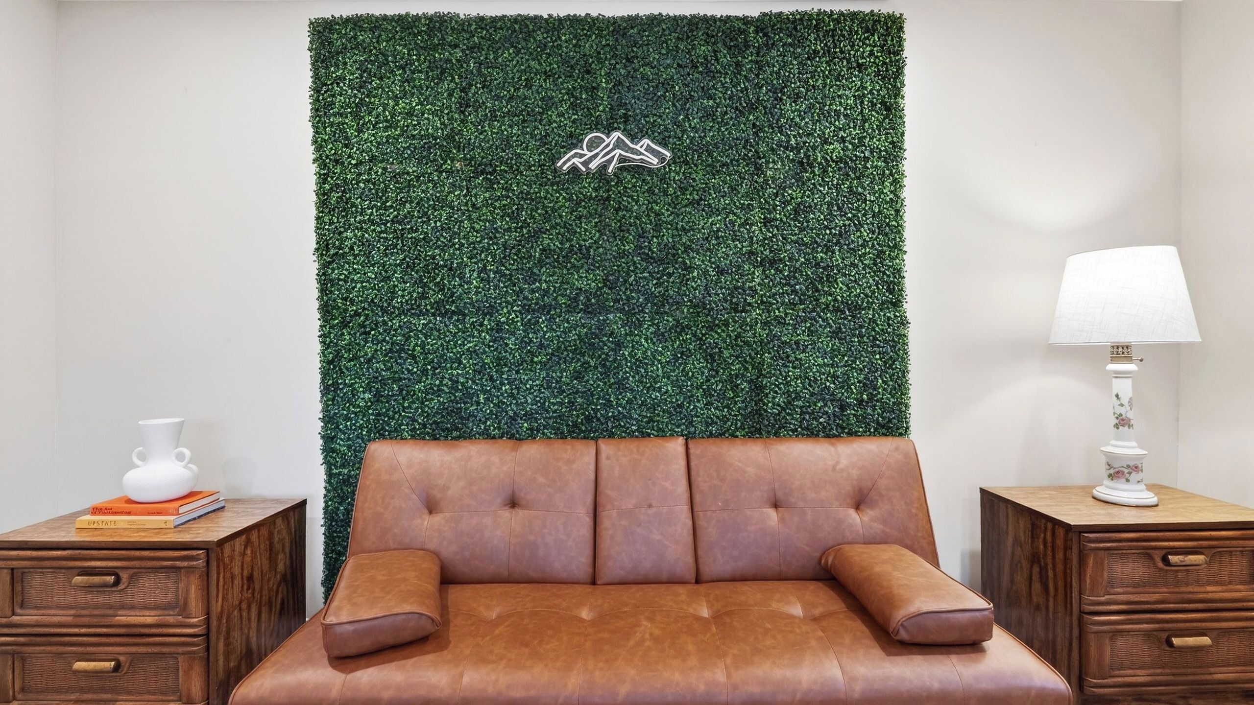 A room with a brown leather sofa, a green wall with a logo, a white vase, and a lamp on wooden nightstands.