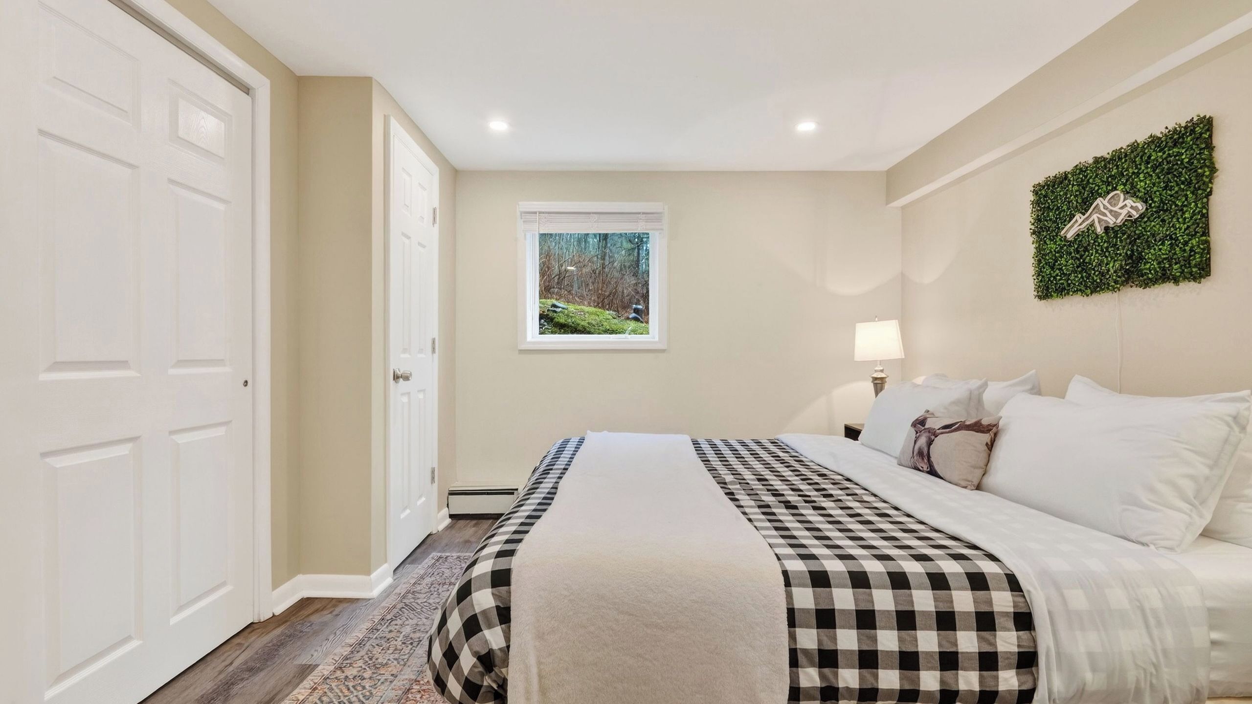 A bedroom with a checkered bedspread, a window with a forest view, a wall-mounted plant, and a door.