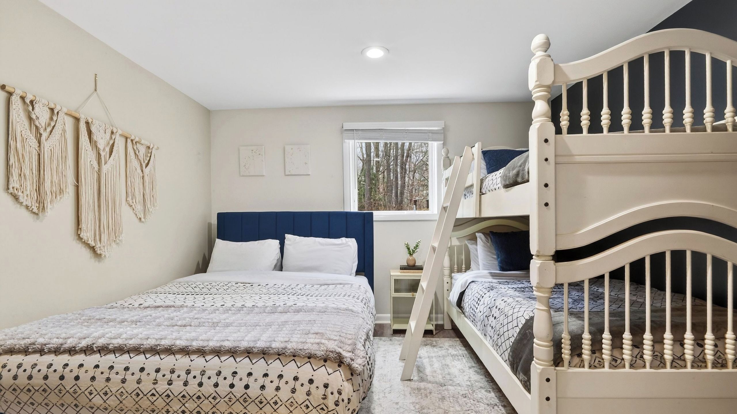 Un dormitorio con una cama litera, una cama individual, adornos de macramé en la pared, una ventana con vistas a árboles y una mesita de noche con una planta.