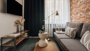 A modern living room with a brick wall, a grey sofa, a coffee table, a TV mounted on the wall, and a floor lamp. - Skawinska Suites by Fragola Apartments (Kraków)