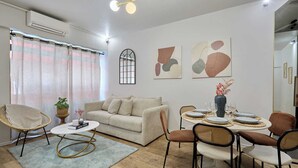 Una sala de estar con sofá, mesa de centro, mesa de comedor con sillas, obra de arte abstracta en la pared y ventana con cortinas.