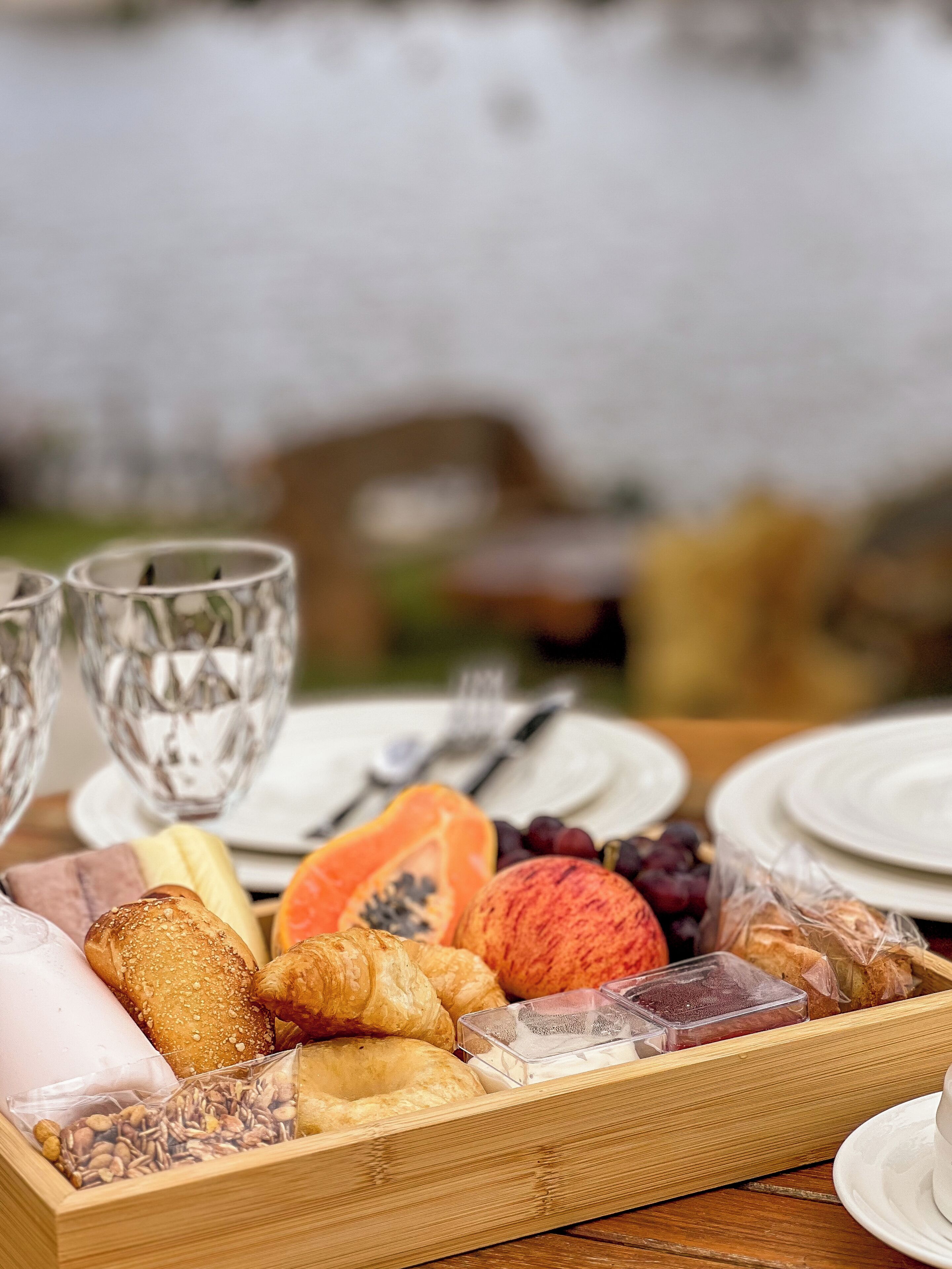 A wooden tray with various food items and a white plate with a cup, set on a table outdoors.