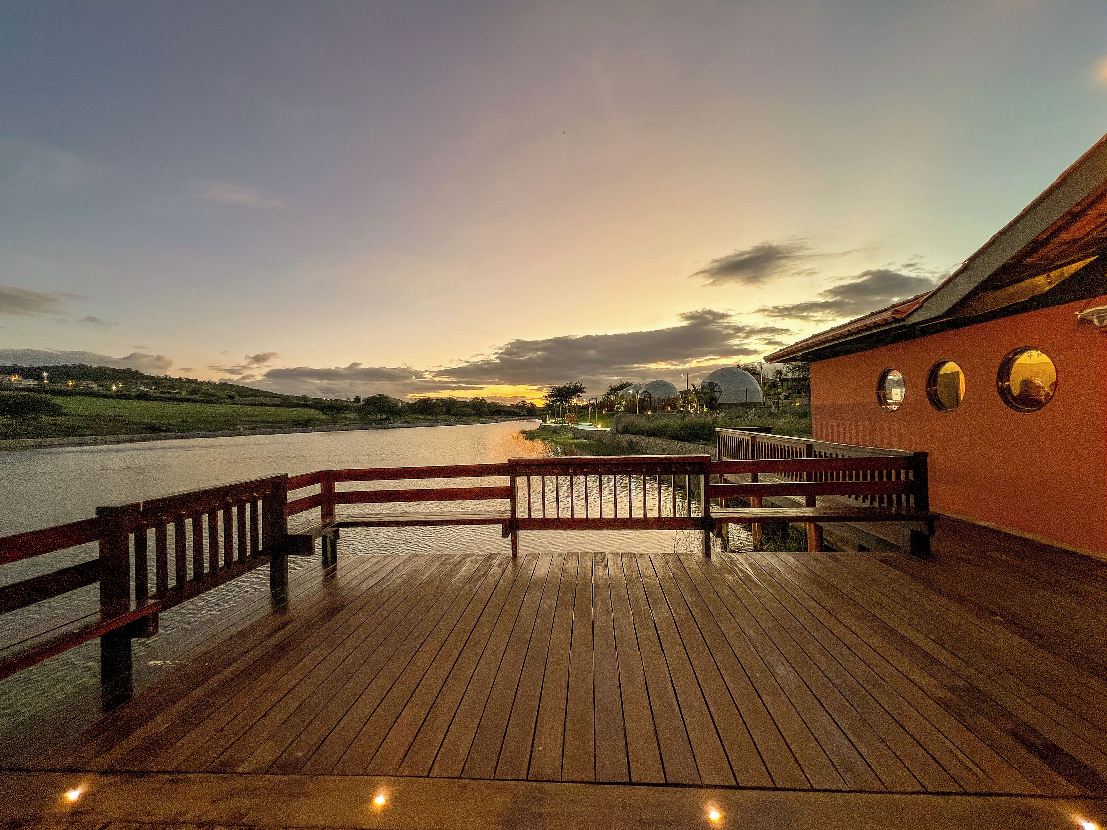 Una terraza de madera con barandilla da vista a una masa de agua, un edificio con tres ventanas circulares y un atardecer al fondo.