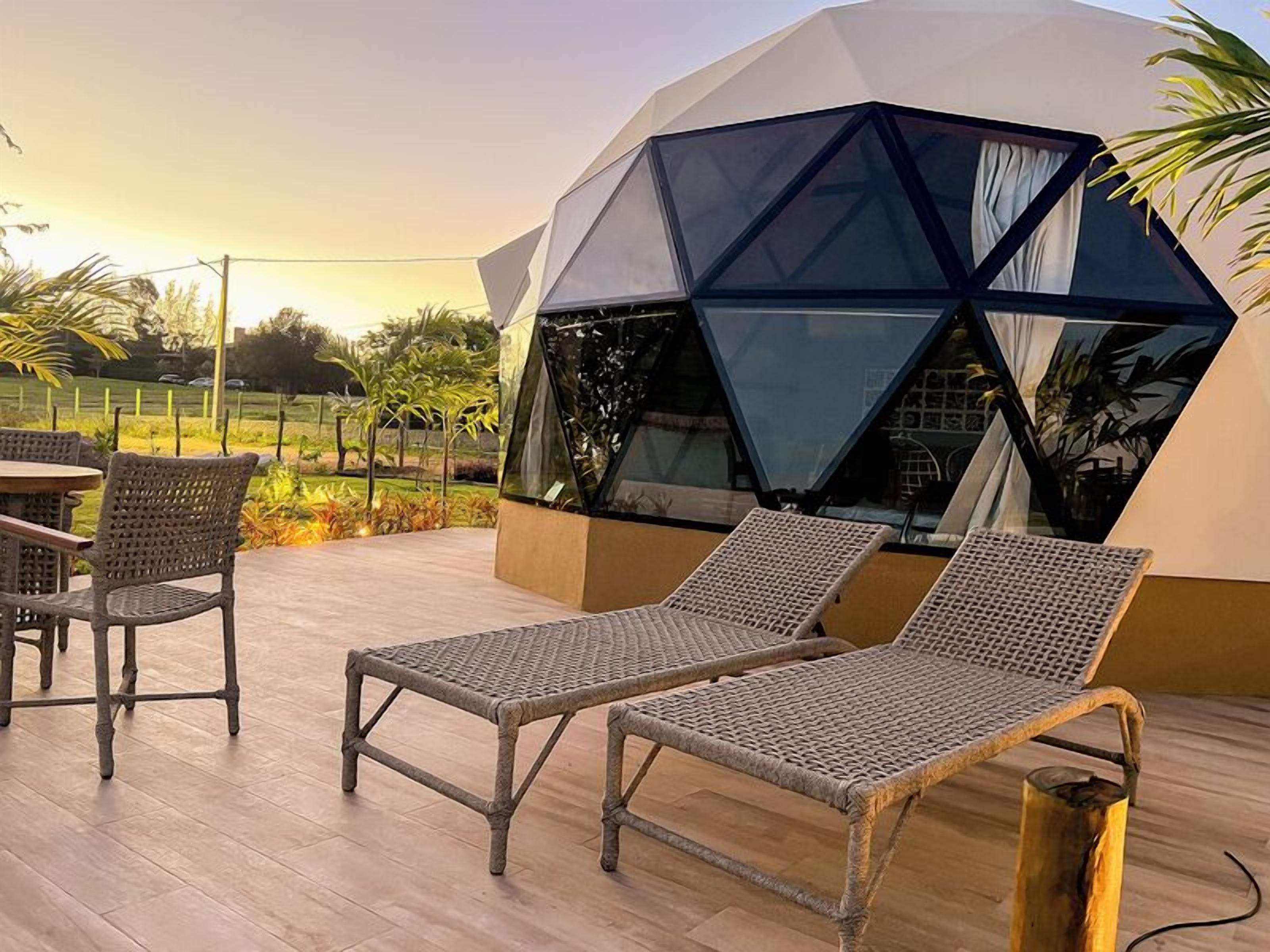Een moderne buitenruimte met loungezetels, een tafel, een geometrische glazen constructie en een houten vloer.